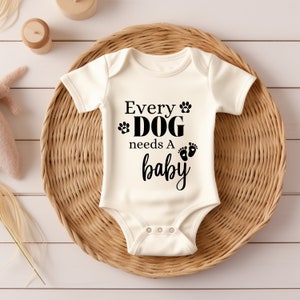 Op de afbeelding: Crème babyromper met de zwarte tekst "Every DOG needs A baby". Het ontwerp bevat pootafdrukken en babyvoetjes. De romper ligt op een geweven rieten dienblad, met een houten speelgoed en een knuffel op de achtergrond.