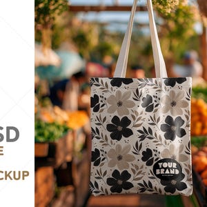 May include: A tote bag mockup with a floral pattern in black, gray, and beige. The bag has white handles and a circular logo that says "YOUR BRAND". The background is blurred, suggesting an outdoor market setting. The text on the left says ".PSD TOTE BAG MOCKUP".