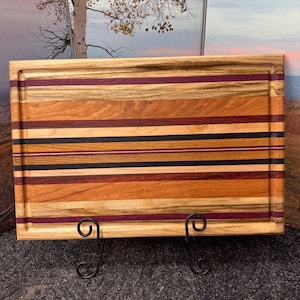 May include: A rectangular wooden cutting board with a juice groove. The board features a striped design with various wood tones, including light brown, dark brown, black, and purple. It is displayed on a black metal stand.