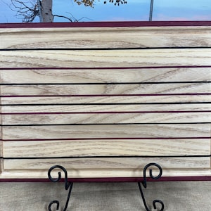 May include: Rectangular wooden cutting board with a light wood grain pattern. The board features horizontal stripes in black and burgundy, and a burgundy border. A groove runs along the inside edge. The board is displayed on a black metal stand.