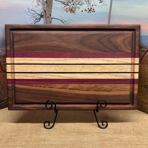 May include: A large rectangular wooden cutting board with a dark brown walnut finish. The board has three stripes of lighter wood and a purple stripe running across the center. The board is on a stand.
