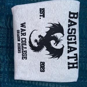 May include: Gray sweatshirt with a black dragon graphic and the text "Basciath War College Dragon Riders Est. 2023"