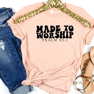 May include: A light pink t-shirt with black text that reads "Made to Worship Psalm 95:1". The text is in a retro font style.