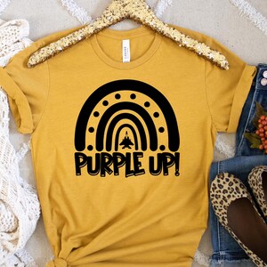 May include: A mustard yellow t-shirt featuring a black silhouette of a rainbow with a jet flying through the centre. The text "Purple Up!" is written below the rainbow.