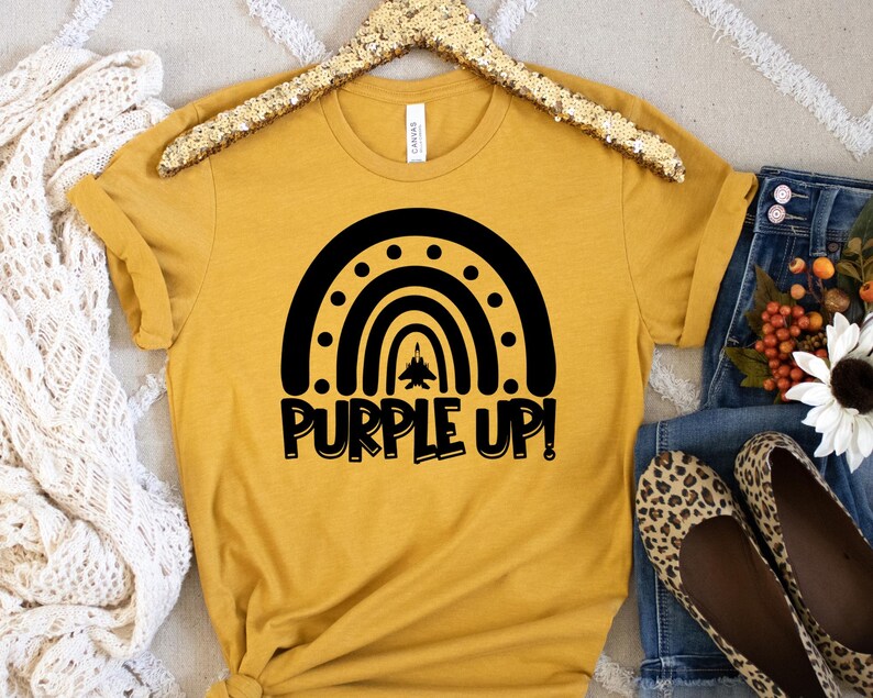May include: A mustard yellow t-shirt featuring a black silhouette of a rainbow with a jet flying through the centre. The text "Purple Up!" is written below the rainbow.