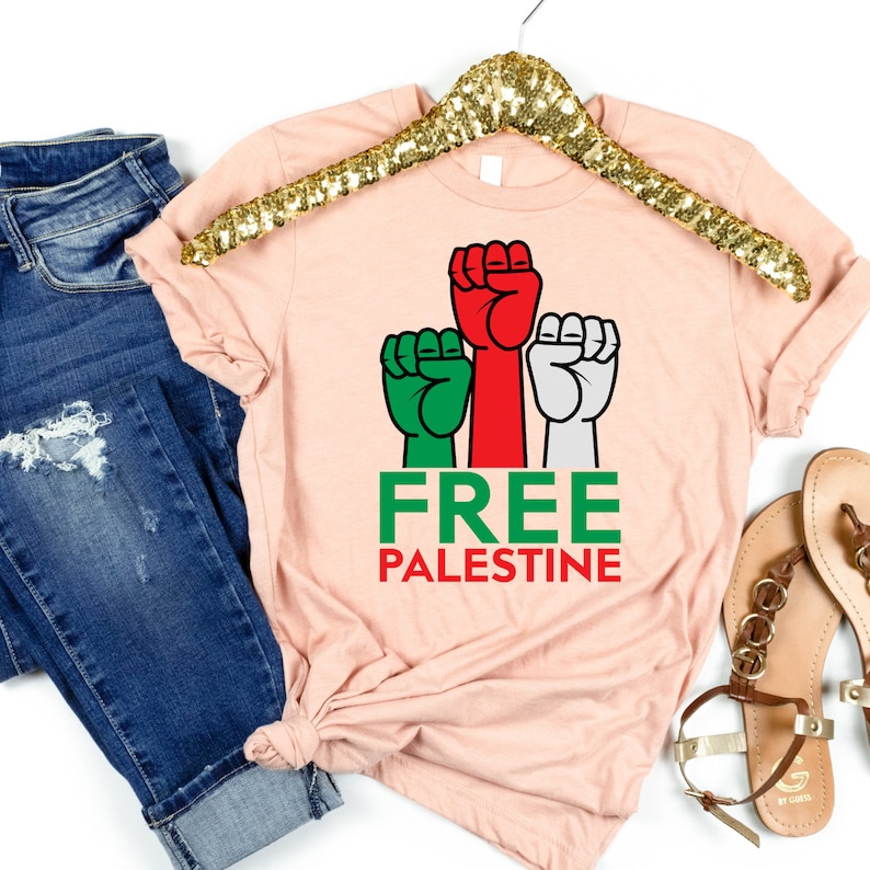 May include: A light pink t-shirt with a graphic of three fists in green, red, and white, with the words "FREE PALESTINE" in green and red. The shirt is hanging on a gold sequined hanger, with blue jeans and sandals in the background.