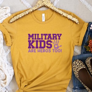 May include: A mustard yellow t-shirt with the text "Military Kids Are Heros Too!" in purple lettering. The shirt has a handprint with a heart in the centre.