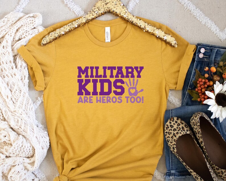 May include: A mustard yellow t-shirt with the text "Military Kids Are Heros Too!" in purple lettering. The shirt has a handprint with a heart in the centre.