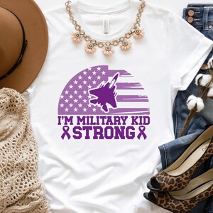 May include: White t-shirt featuring a purple silhouette of a fighter jet flying over a purple and white American flag. The text "I'm Military Kid Strong" is printed in purple below the image.