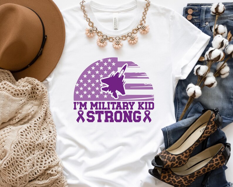 May include: White t-shirt featuring a purple silhouette of a fighter jet flying over a purple and white American flag. The text "I'm Military Kid Strong" is printed in purple below the image.