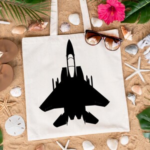 May include: A white canvas tote bag featuring a black silhouette of a fighter jet printed on it. The bag is lying on a sandy surface with seashells, starfish, and tropical leaves.