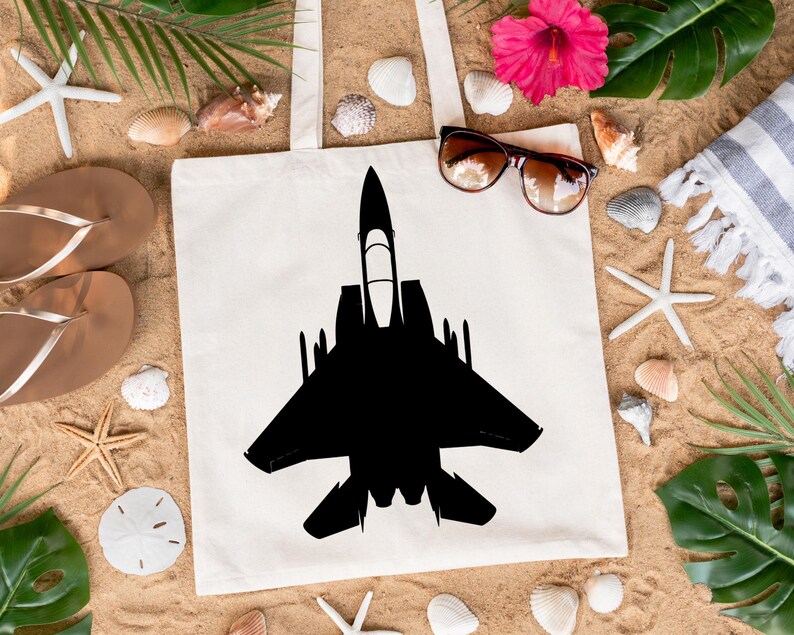 May include: A white canvas tote bag featuring a black silhouette of a fighter jet printed on it. The bag is lying on a sandy surface with seashells, starfish, and tropical leaves.