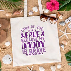 May include: White canvas tote bag with purple text that reads "Land of the Free Because My Daddy Is Brave #Military Child". The bag is lying on a sandy beach with seashells, starfish, and green leaves.