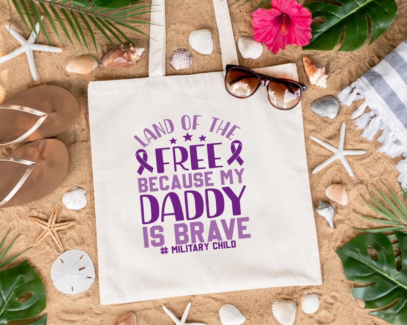 May include: White canvas tote bag with purple text that reads "Land of the Free Because My Daddy Is Brave #Military Child". The bag is lying on a sandy beach with seashells, starfish, and green leaves.