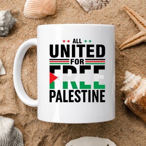 May include: White ceramic mug with the text "ALL UNITED FOR FREE PALESTINE" in black, green, red, and white. The mug features the Palestinian flag colors and is set against a sandy background with seashells.