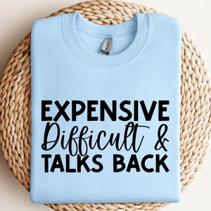 Könnte beinhalten: Hellblauer Pullover mit dem Text "Expensive Difficult & Talks Back" in Schwarz.