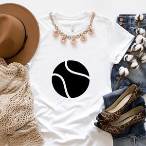 May include: White t-shirt with a black tennis ball graphic. The shirt is styled with a brown hat, a necklace, jeans, and leopard print shoes.
