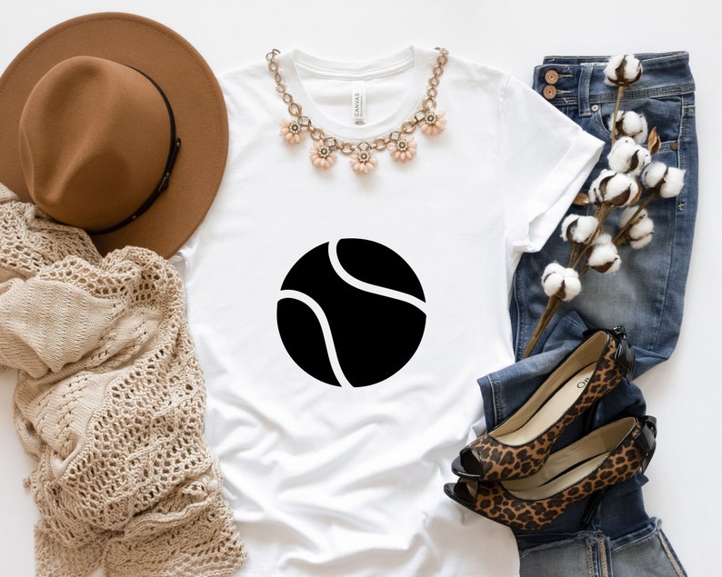 May include: White t-shirt with a black tennis ball graphic. The shirt is styled with a brown hat, a necklace, jeans, and leopard print shoes.