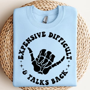 Può includere: Felpa blu chiaro con una grafica nera di una mano scheletrica che fa il segno shaka. La grafica è circondata da un cerchio con il testo "Expensive, Difficult & Talks Back" in un font retrò.