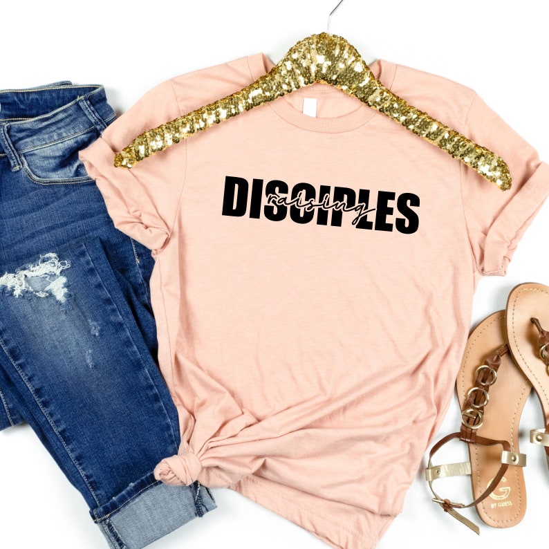 May include: A peach-colored t-shirt with black text that reads "raising disciples". The shirt is on a white background with a gold hanger, blue jeans, and brown sandals.
