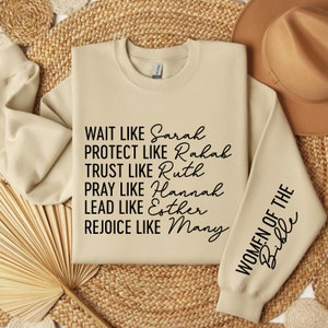 May include: A beige sweatshirt with black text that reads "WAIT LIKE Sarah PROTECT LIKE Rahah TRUST LIKE Ruth PRAY LIKE Hannah LEAD LIKE Esther REJOICE LIKE Many" on the front and "WOMEN OF THE Bible" on the sleeve.