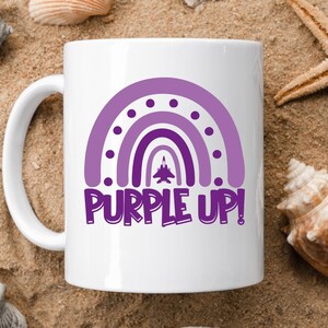 May include: White ceramic mug featuring a purple rainbow design with the text "PURPLE UP!" printed on it. A small purple airplane is flying through the rainbow.