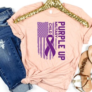 May include: Purple Up for Military Child t-shirt featuring a purple ribbon and an American flag design. The shirt is a light pink colour.