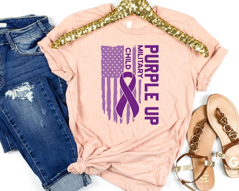 May include: Purple Up for Military Child t-shirt featuring a purple ribbon and an American flag design. The shirt is a light pink colour.