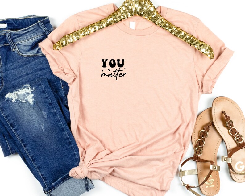 May include: A light pink t-shirt with the text "YOU matter" in black lettering on the front. The shirt is folded and displayed on a white background with a pair of sandals and blue jeans.