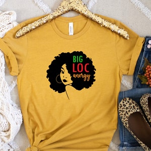 May include: A mustard yellow t-shirt featuring a black silhouette of a woman with large afro hair. The text "BIG LOC energy" is written in green, red, and black letters.