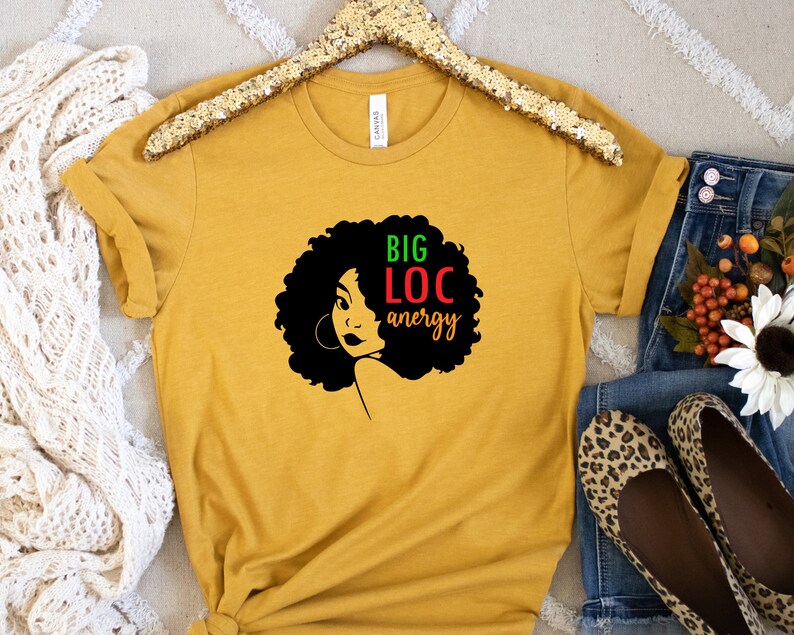 May include: A mustard yellow t-shirt featuring a black silhouette of a woman with large afro hair. The text "BIG LOC energy" is written in green, red, and black letters.