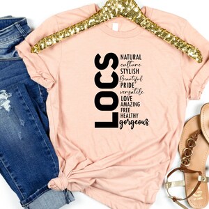 May include: A peach-coloured t-shirt with the word "LOCS" in black vertical letters. The letters are surrounded by words describing the characteristics of locs, including "natural", "culture", "stylish", "beautiful", "pride", "versatile", "love", "amazing", "free", "healthy", and "gorgeous".