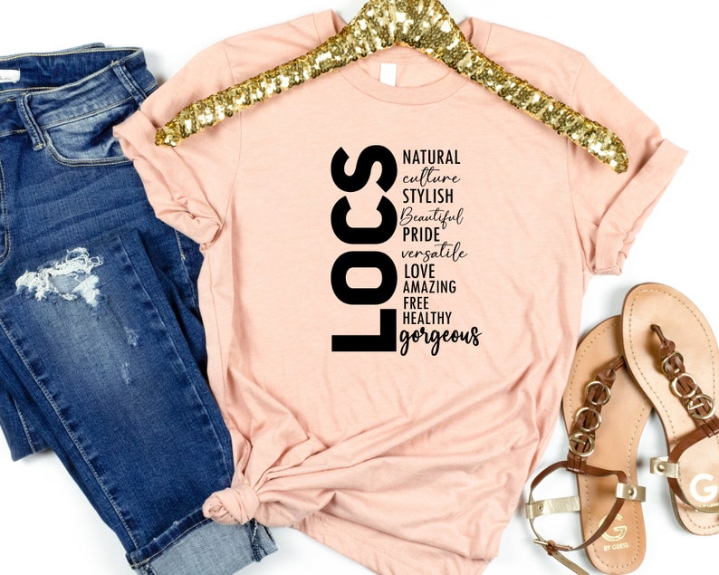 May include: A peach-coloured t-shirt with the word "LOCS" in black vertical letters. The letters are surrounded by words describing the characteristics of locs, including "natural", "culture", "stylish", "beautiful", "pride", "versatile", "love", "amazing", "free", "healthy", and "gorgeous".