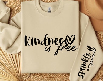 Può includere: Una felpa beige con la scritta "kindness is free" e un cuore sul davanti. La manica sinistra ha la scritta "sprinkle it everywhere" in nero.