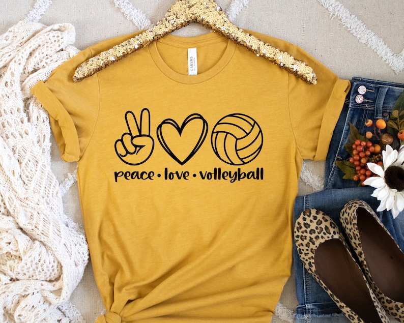 K&ouml;nnte beinhalten: Ein senfgelbes T-Shirt mit einem Peace-Zeichen, einem Herzen und einer Volleyball-Grafik. Der Text "peace love volleyball" ist unter der Grafik aufgedruckt.
