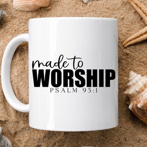 May include: White ceramic mug with black text that reads "made to worship" and "PSALM 95:1". The mug is sitting on a sandy surface with seashells and a starfish.