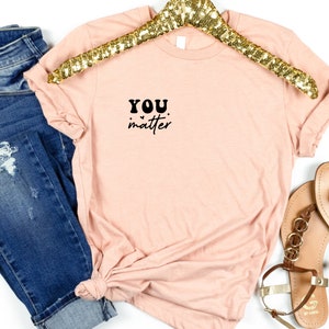 May include: A light pink t-shirt with the text "YOU matter" in black lettering on the front. The shirt is folded and displayed on a white background with a gold sequined hanger and brown sandals.