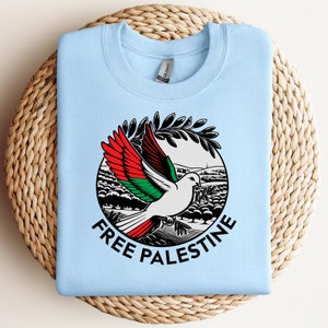 May include: Light blue sweatshirt featuring a dove with wings in the colors of the Palestinian flag, encircled by an olive branch and a landscape. The words "FREE PALESTINE" are printed in a semi-circular design below the dove.