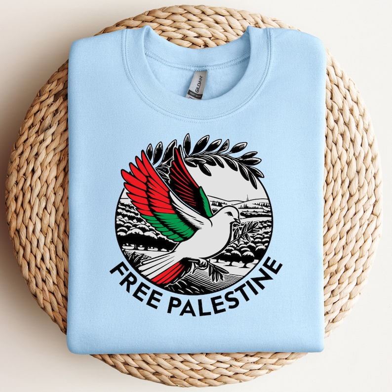 May include: Light blue sweatshirt featuring a dove with wings in the colors of the Palestinian flag, encircled by an olive branch and a landscape. The words "FREE PALESTINE" are printed in a semi-circular design below the dove.