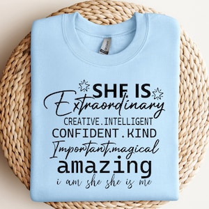 Op de afbeelding: Lichtblauw sweatshirt met zwarte tekst die luidt: "Zij is buitengewoon. Creatief. Intelligent. Zelfverzekerd. Aardig. Belangrijk. Magisch. Geweldig. Ik ben zij, zij is ik."
