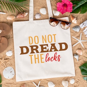 May include: A white tote bag with the text "Do Not Dread The Locks" in brown, yellow, and red letters. The bag is surrounded by seashells, starfish, and tropical plants on a sandy beach.