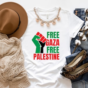 May include: White t-shirt with the words "FREE GAZA FREE PALESTINE" in green and red, and a black fist graphic. The shirt is styled with a brown hat, a beige sweater, a necklace, jeans, and leopard print heels.