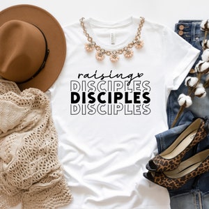 May include: White t-shirt with the text "raising DISCIPLES DISCIPLES DISCIPLES" in black. The shirt is laid flat on a white surface with a brown hat, a beige knitted scarf, a necklace with pink flower charms, blue jeans, and a pair of leopard print heels.