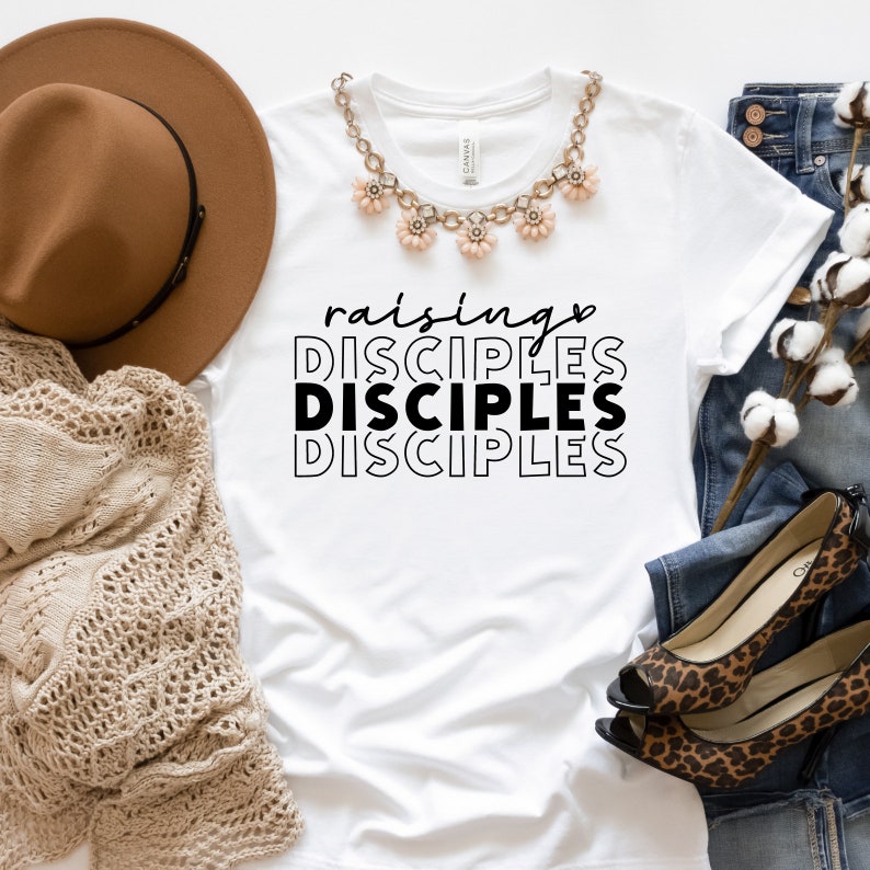 May include: White t-shirt with the text "raising DISCIPLES DISCIPLES DISCIPLES" in black. The shirt is laid flat on a white surface with a brown hat, a beige knitted scarf, a necklace with pink flower charms, blue jeans, and a pair of leopard print heels.