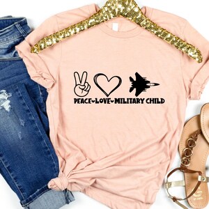 May include: A pink t-shirt with a peace sign, heart, and jet graphic. The text "Peace-Love-Military Child" is printed on the shirt.