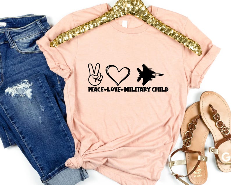 May include: A pink t-shirt with a peace sign, heart, and jet graphic. The text "Peace-Love-Military Child" is printed on the shirt.