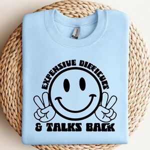 Op de afbeelding: Lichtblauw sweatshirt met zwarte tekst die "Duur, Moeilijk & Praat Terug" zegt met een smiley en peace-tekens.