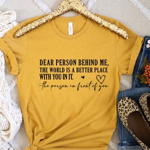 May include: A mustard yellow t-shirt with the text "DEAR PERSON BEHIND ME, THE WORLD IS A BETTER PLACE WITH YOU IN IT. -the person in front of you" printed in black.