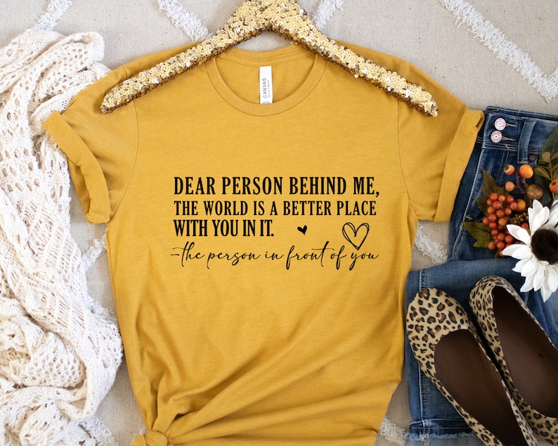 May include: A mustard yellow t-shirt with the text "DEAR PERSON BEHIND ME, THE WORLD IS A BETTER PLACE WITH YOU IN IT. -the person in front of you" printed in black.