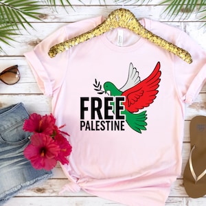May include: Light pink t-shirt with a dove graphic in green, white, and red, holding an olive branch. The words "FREE PALESTINE" are printed in black, with the letter "E" in green. The shirt is displayed on a gold hanger.
